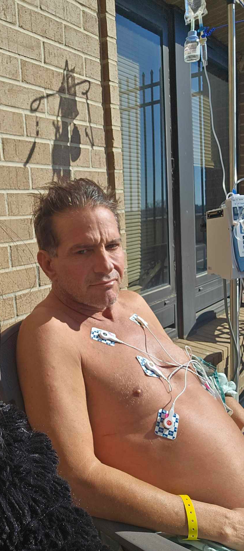 David recovering on the sixth-floor hospital balcony in February 2024, wearing heart monitor electrodes with an IV pole beside him