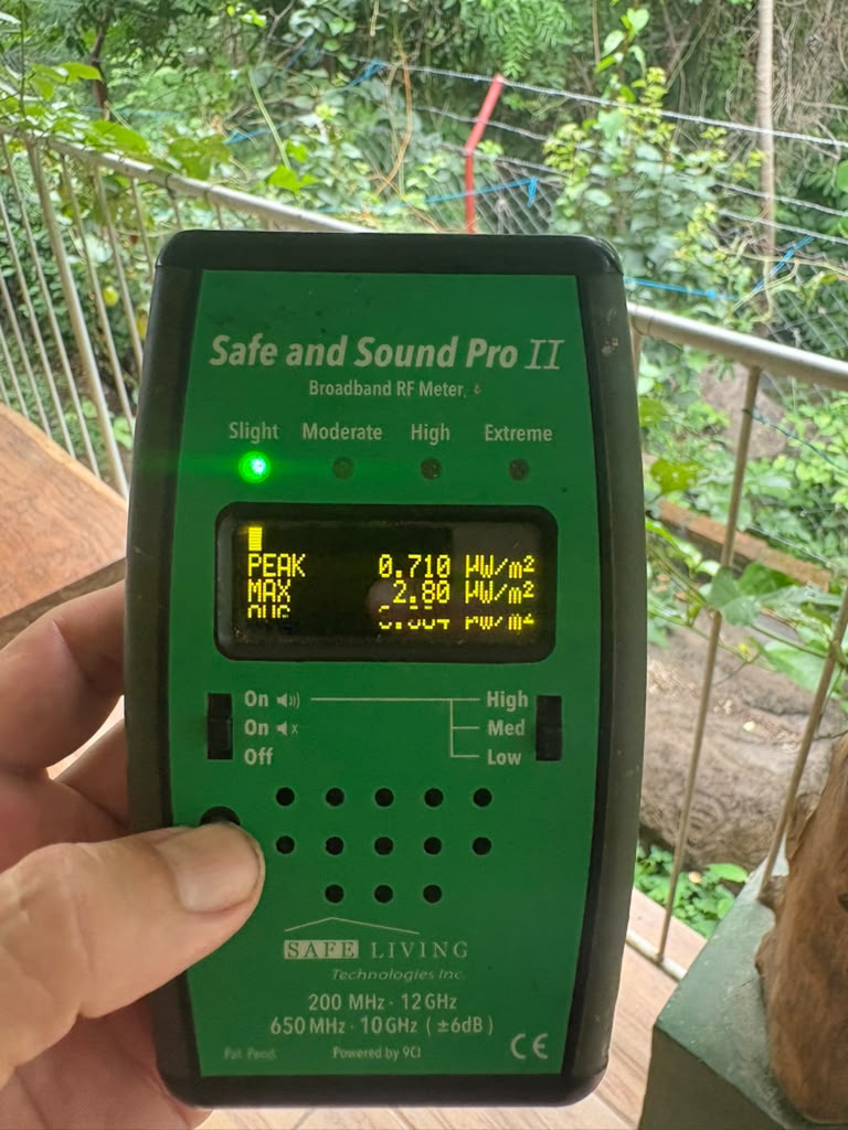 A hand holding a Safe and Sound Pro II broadband RF meter on a porch in Ometepe, showing PEAK 0.710 and MAX 2.80 µW/m²