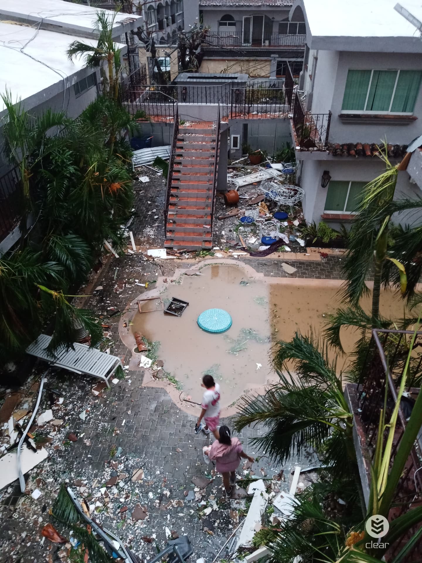 The same Acapulco courtyard after Hurricane Otis, with debris scattered across the ground and the pool filled with muddy floodwater